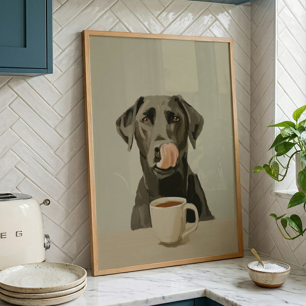 A framed art print of a black dog with its tongue out licking a white coffee mug, displayed on a kitchen counter against a white herringbone tile backsplash.