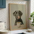 A framed art print of a black dog with its tongue out licking a white coffee mug, displayed on a kitchen counter against a white herringbone tile backsplash.