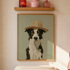 A framed art print of a black and white dog wearing a straw hat, displayed on a bathroom wall above a toilet tank with a wooden shelf holding decorative items.