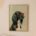 Minimalist oil painting print of a black and white cocker spaniel in side profile wearing gold rimmed amber round sunglasses, painted in rich monochrome tones against a soft sage green background